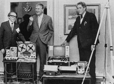 Schwarz-Weiß-Foto von drei Männern neben Tonbandgeräten und Audiotechnik